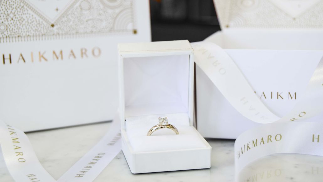 anillo para proponer matrimonio de oro 18k amarillo con un diamante Haikmaro Anillo de compromiso de oro 18k amarillo con diamante colocado en una caja blanca sobre una superficie de mármol. . A su alrededor se ven cintas y empaques de la marca “HAIKMARO” en tonos blancos y dorados